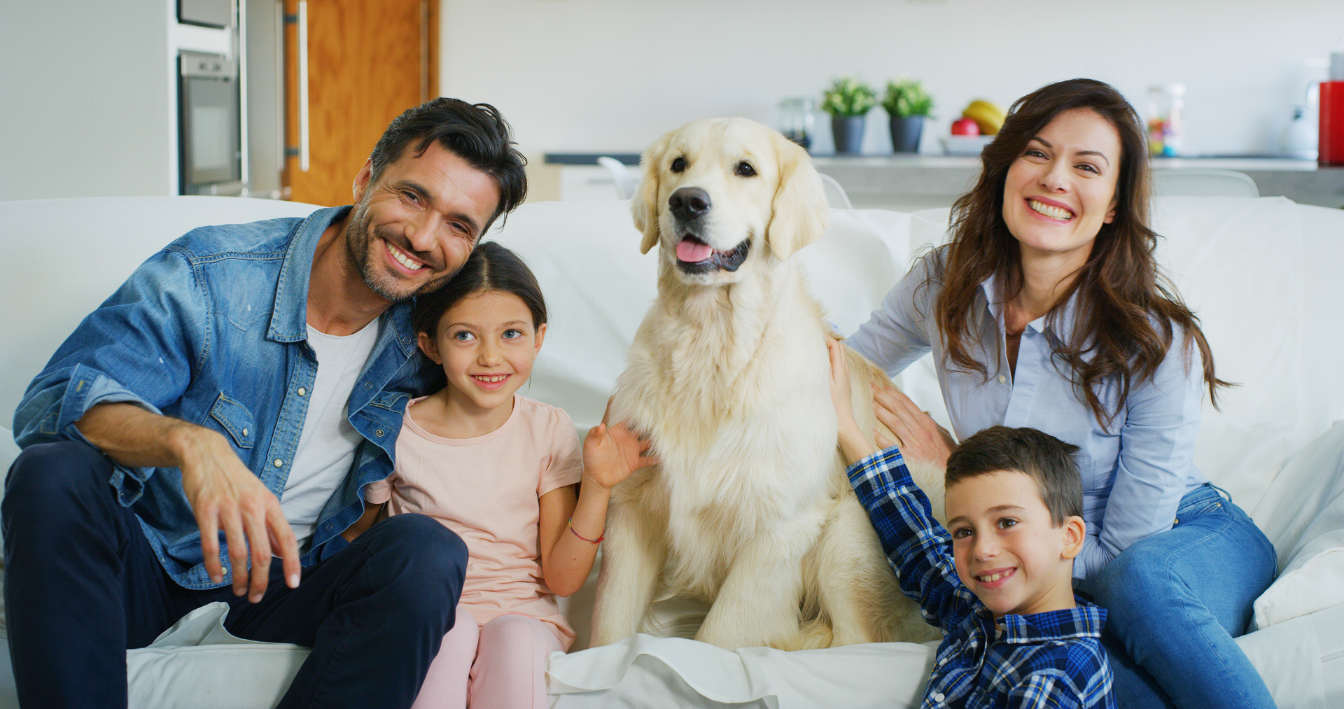 Family of four with a golden retriever dog smiles on a white couch.