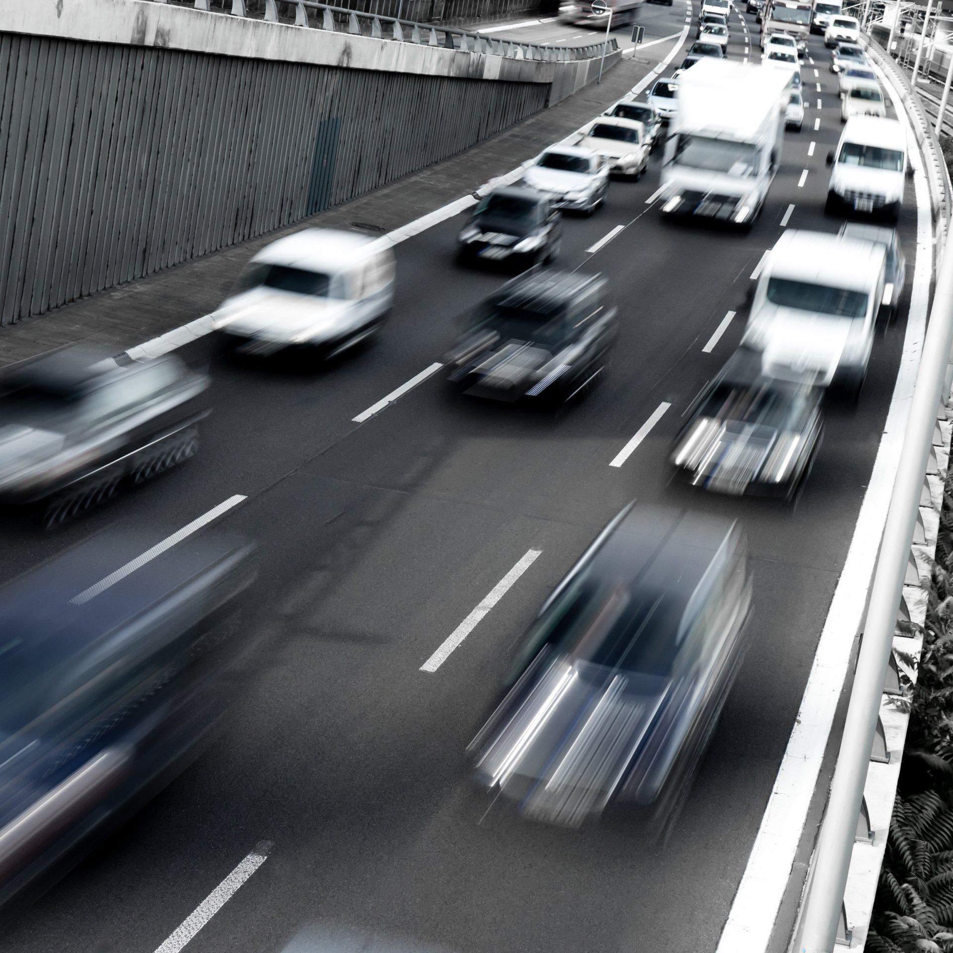 Cars blurred in motion on a multilane highway, beside a concrete retaining wall, overcast sky.