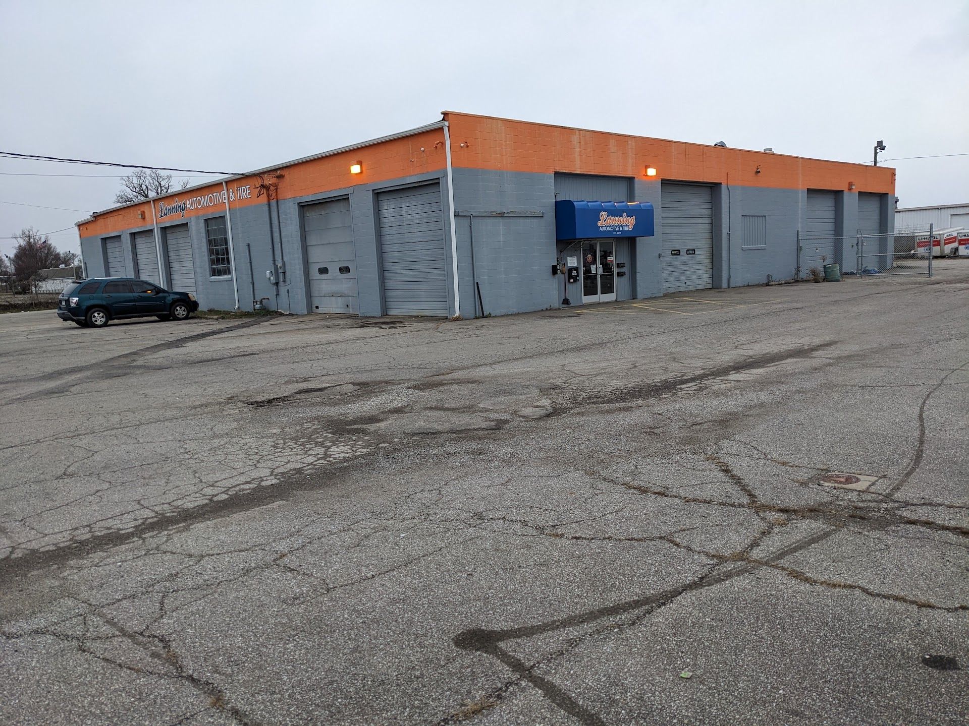 Gray industrial building with orange trim and a blue sign, seen across an empty cracked parking lot | Lanning Automotive & Tire