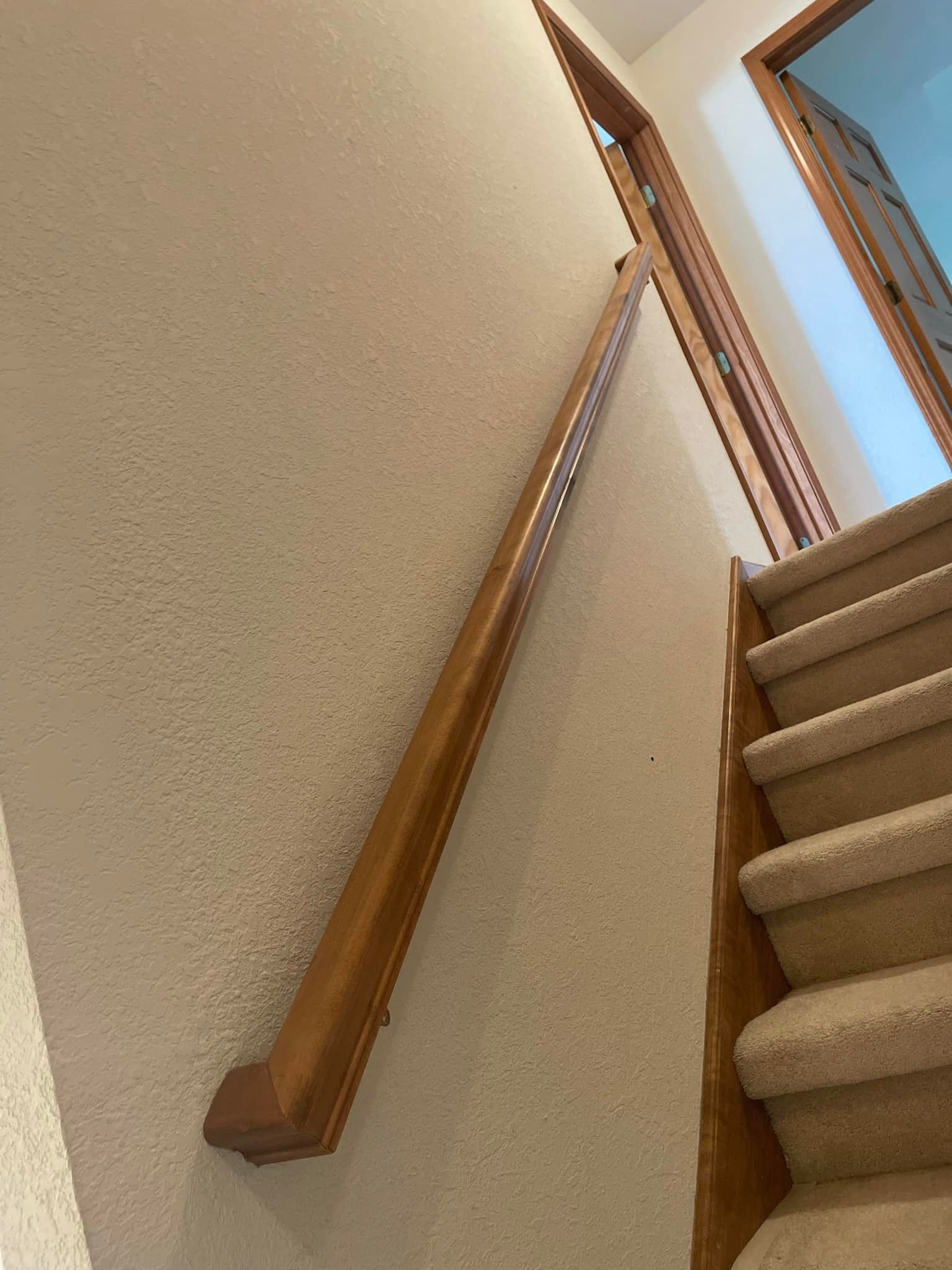 Wooden handrail on a beige wall alongside carpeted stairs.
