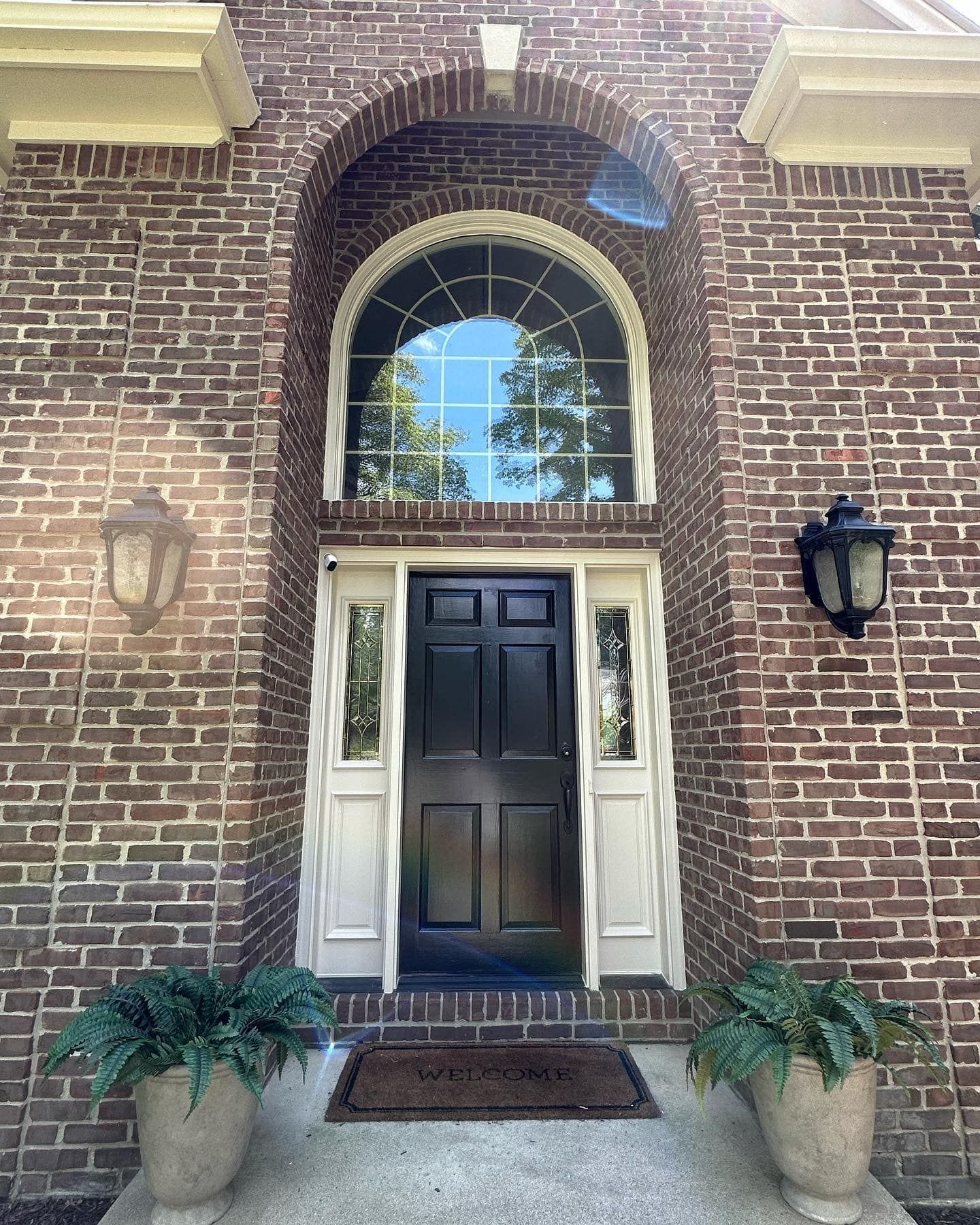 Brick facade with arched window above a black door, flanked by sidelights and sconces, potted plants below.