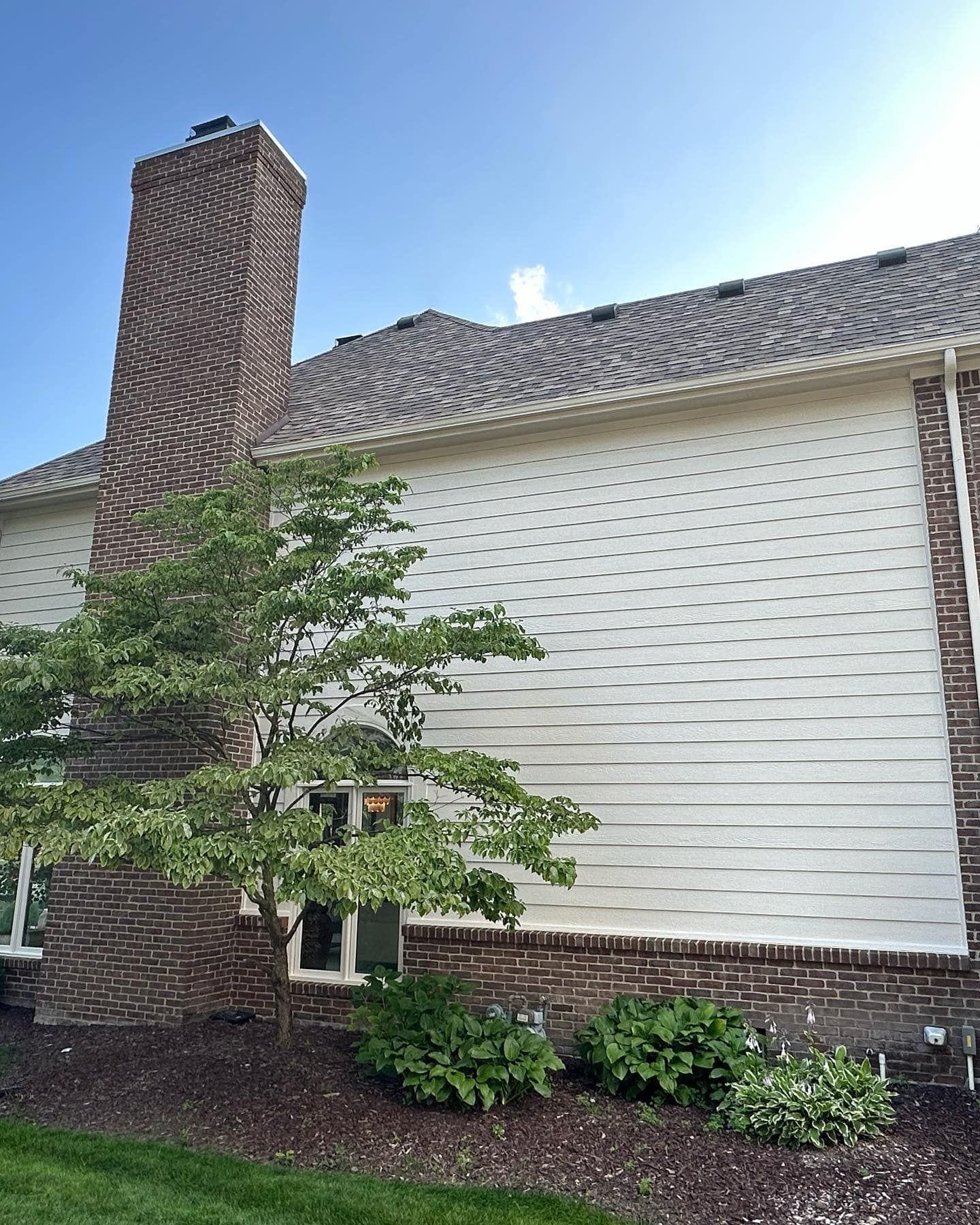 Side of a house with brick chimney, white siding, small tree, and shrubs against a blue sky.
