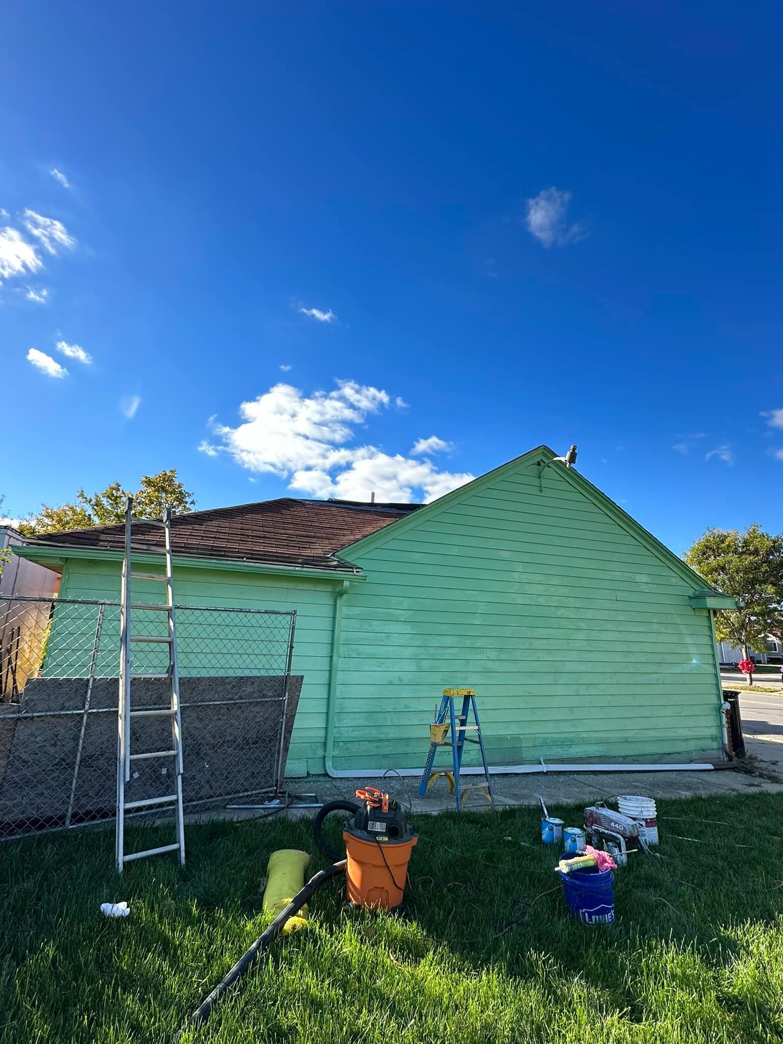 Green-painted building with tools on the grass, ladder, blue sky, and a chain link fence.