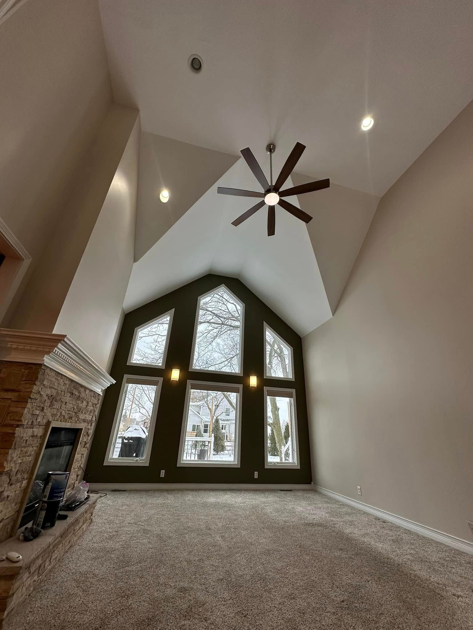 High-ceiling living room with a large window, fireplace, and ceiling fan. White walls, beige carpet.