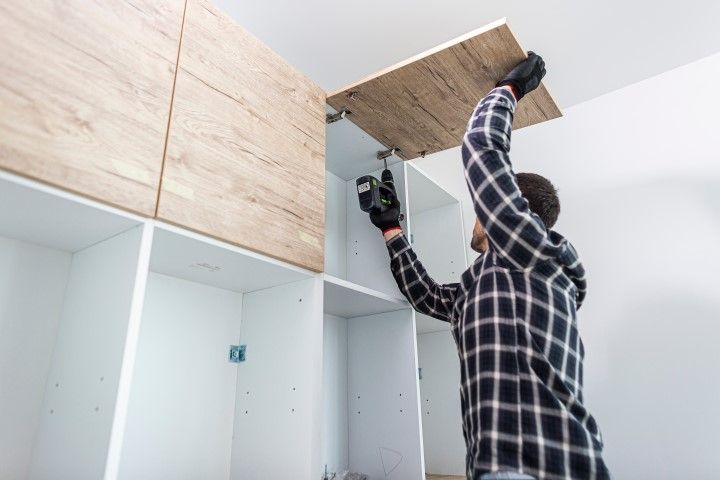 Person installing a wooden cabinet panel with a power drill in a white kitchen.