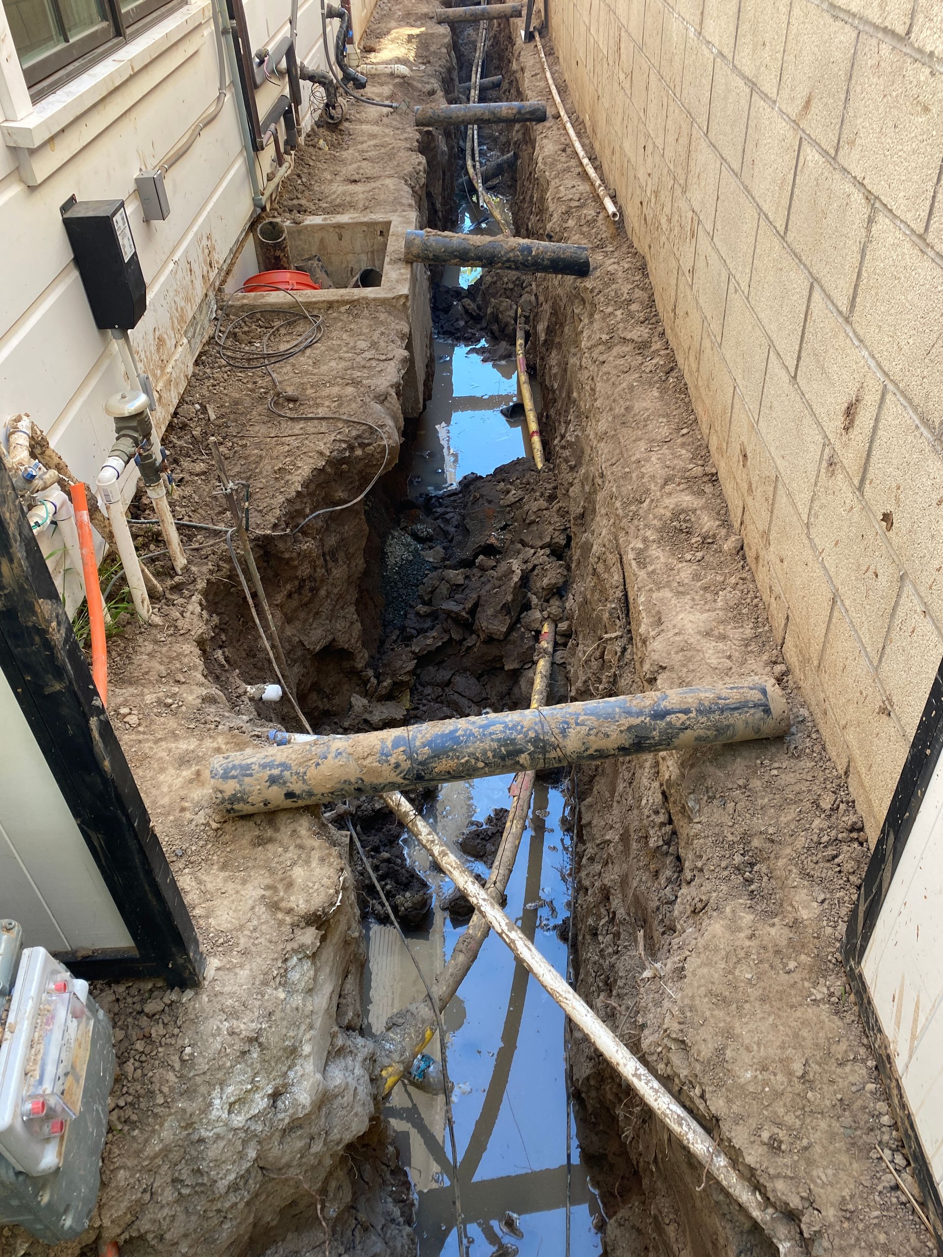 Deep trench between buildings with muddy walls, standing water, and wooden support beams for construction work