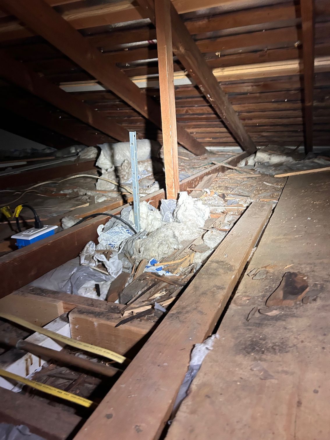 Damaged attic floor with exposed insulation, broken boards, and low wooden beams.