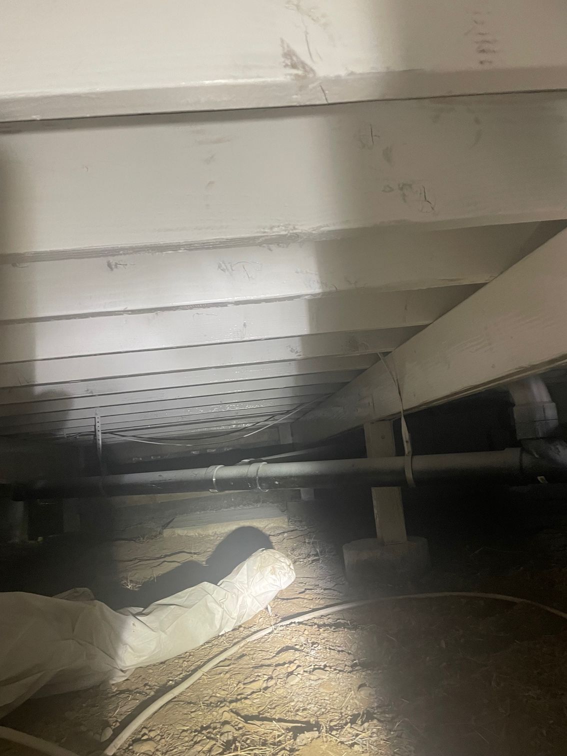 Muddy crawlspace under floor joists with exposed pipes and a bright flashlight beam on the ground