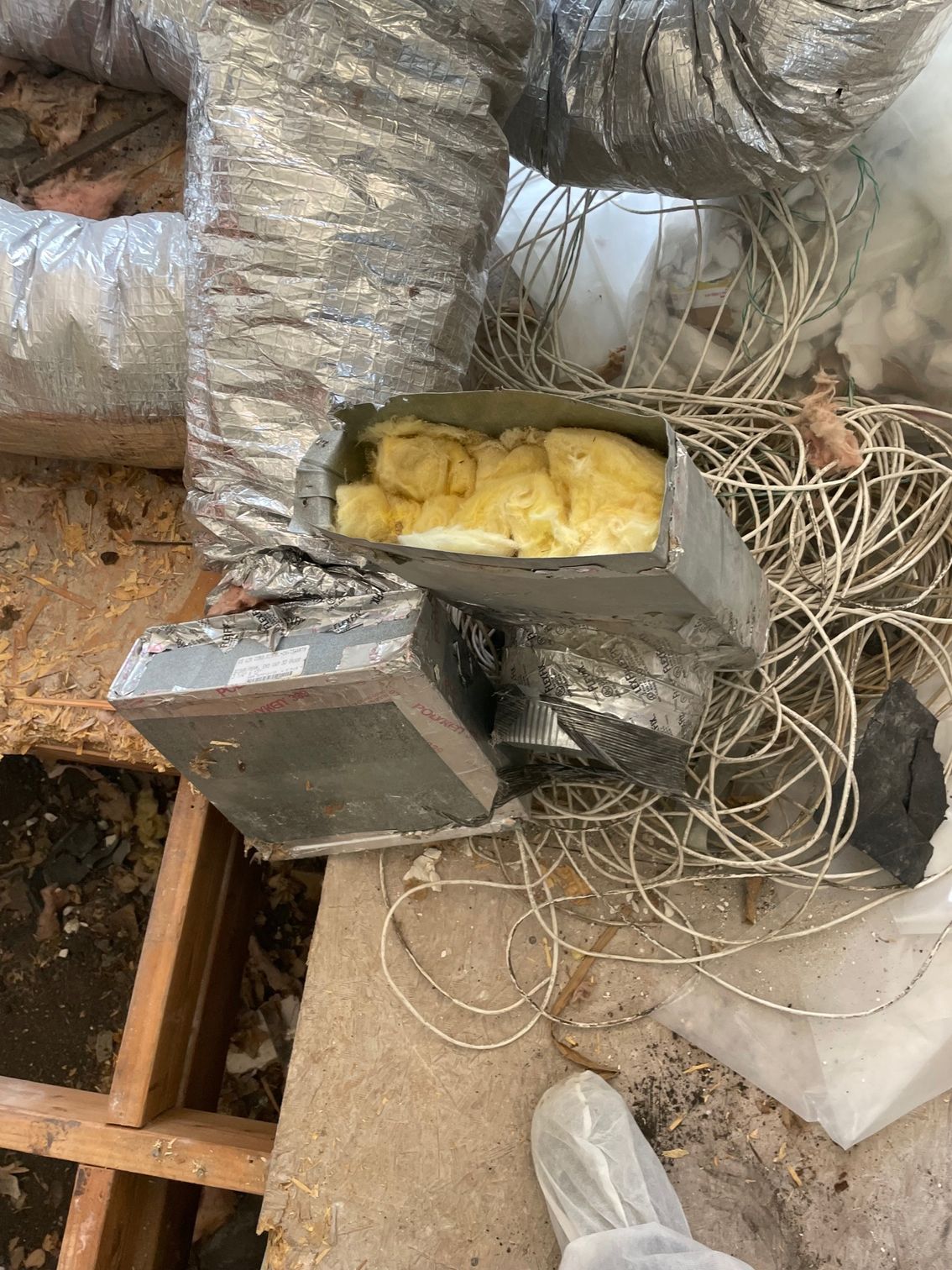 Open HVAC duct with yellow insulation in an unfinished attic, surrounded by silver insulated vents and debris
