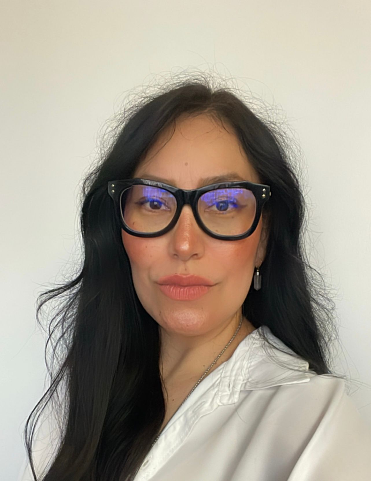 Woman with long black hair wearing oversized black glasses and a white blouse against a plain wall.