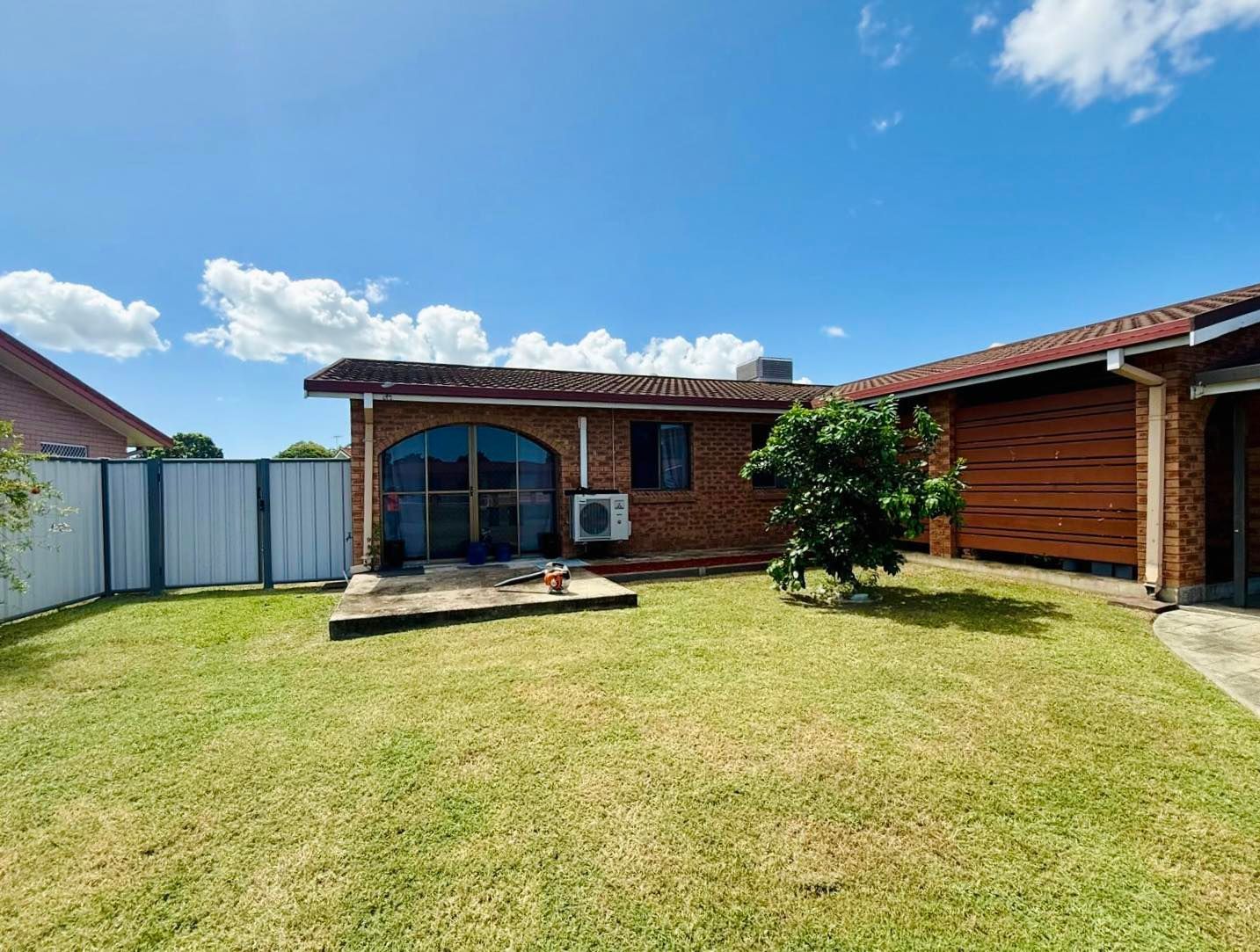 Grassy Backyard With a Red Brick House Under a Partly Cloudy Blue Sky — Hanta Mowing Services Townsville in Kirwan, QLD