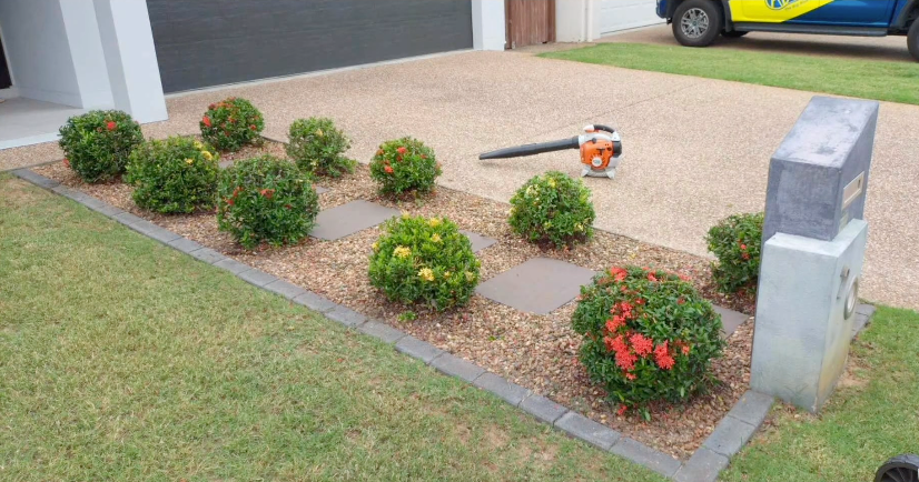 Front yard garden bed with round shrubs, red flowers, and an orange trimmer near a driveway — Hanta Mowing Services Townsville in Kirwan, QLD