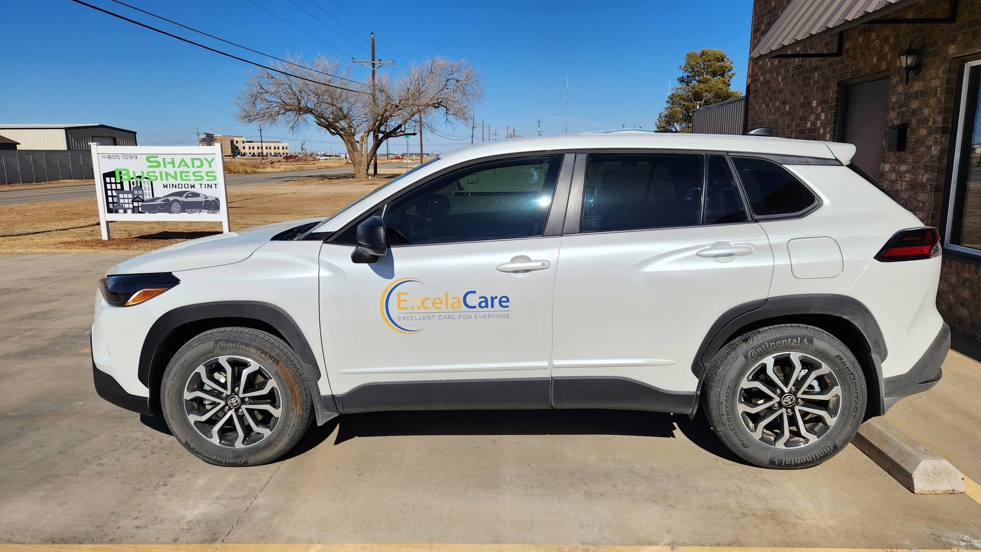 White SUV with “Good Care” logo parked beside a sign on a sunny driveway