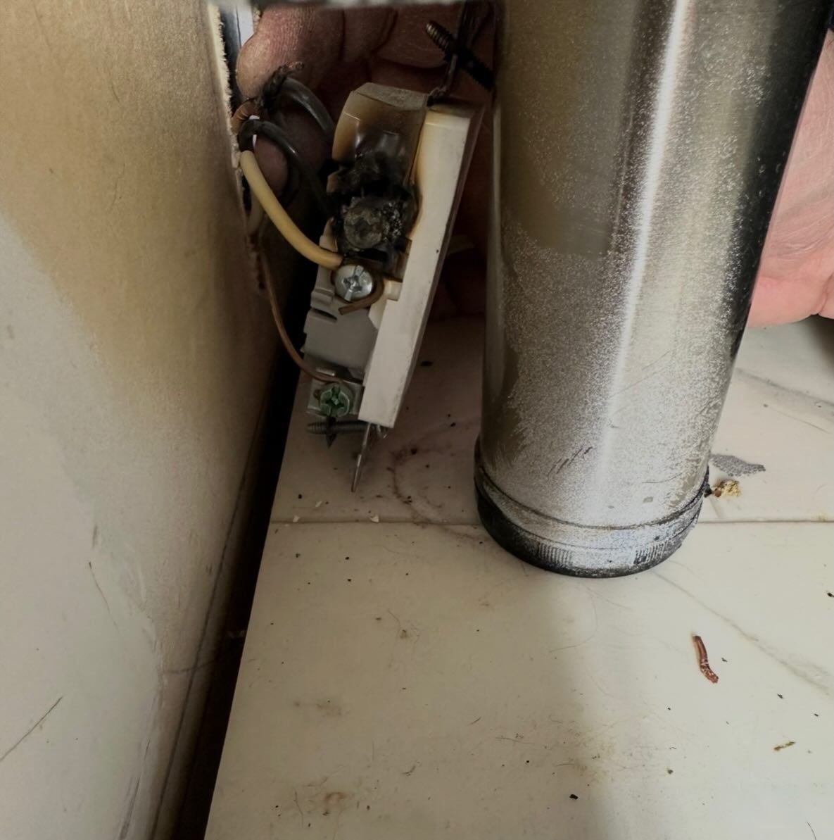 Small electrical component tucked beside a silver duct on a dusty floor near a wall