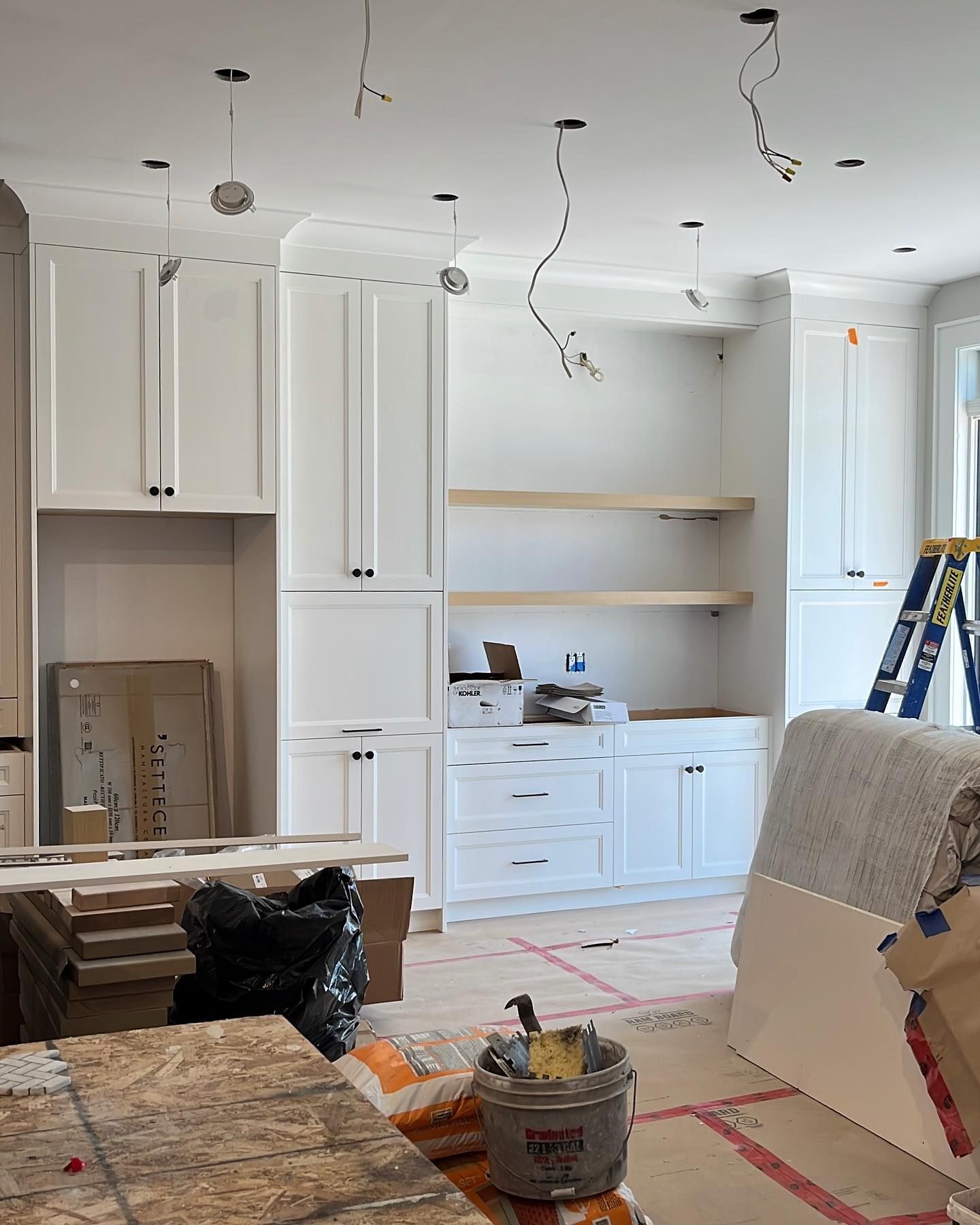 Partially renovated kitchen with white cabinets, open shelving, exposed ceiling wires, and construction materials