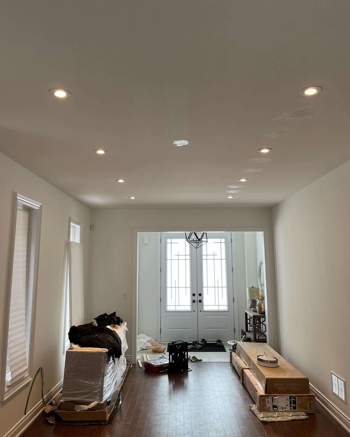 Long, empty living room with white walls, recessed lights, wood floor, and boxes near the far window.