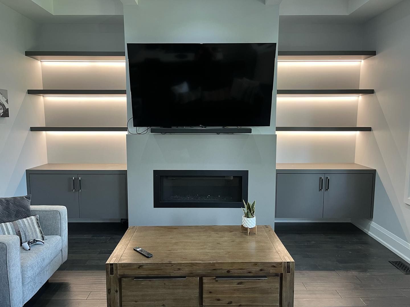 Modern living room with TV above fireplace, built-in shelves, gray cabinets, and a wooden coffee table