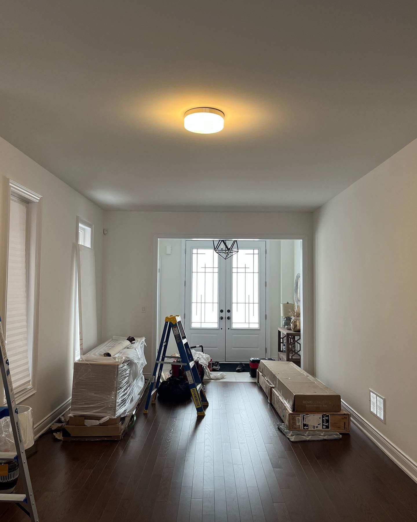 Empty living room with dark wood floors, boxes and tools, and a bright window at the far end.