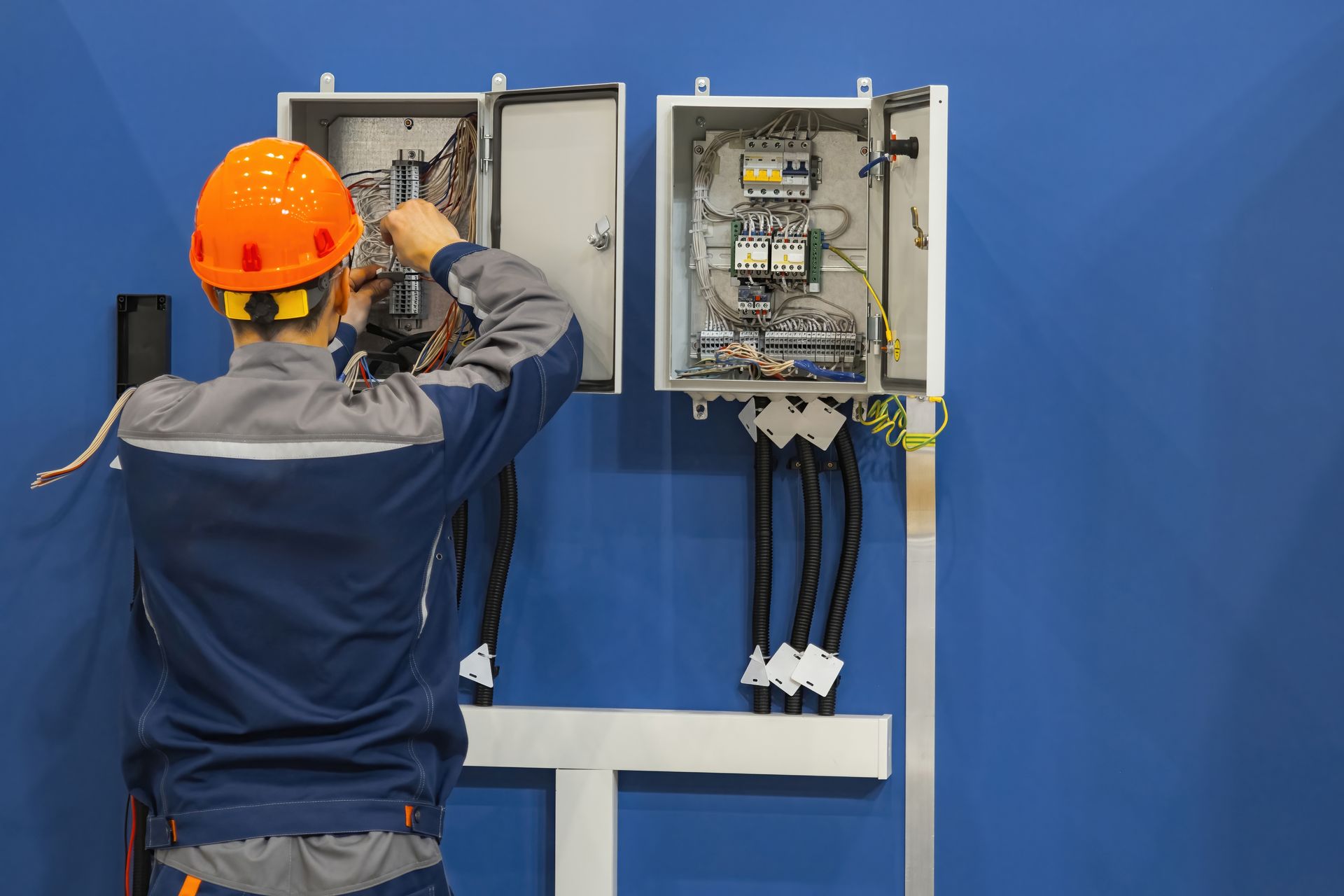 Worker in orange hard hat wiring an open electrical panel against a blue wall.