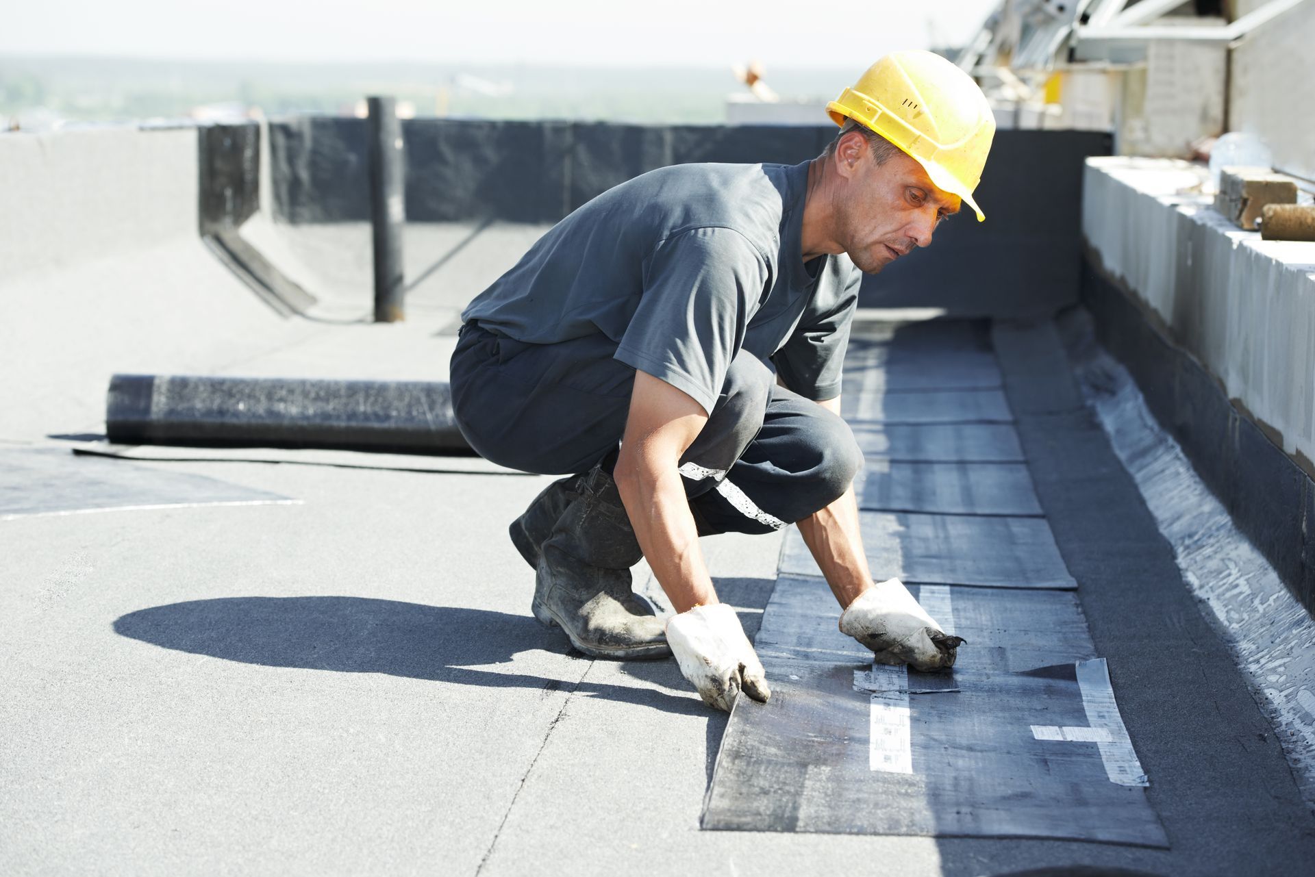 Roofing specialist in a hard hat installing a rolled roofing membrane on a flat roof.