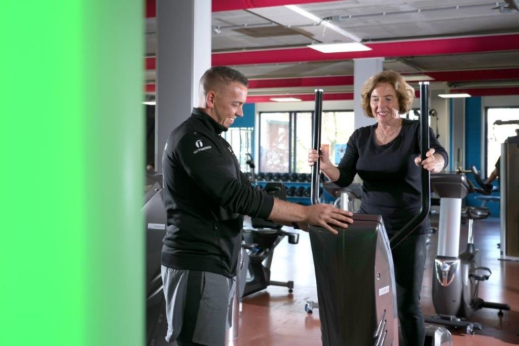 Man helpt vrouw met het gebruiken van een crosstrainer in de sportschool; beiden lachen.