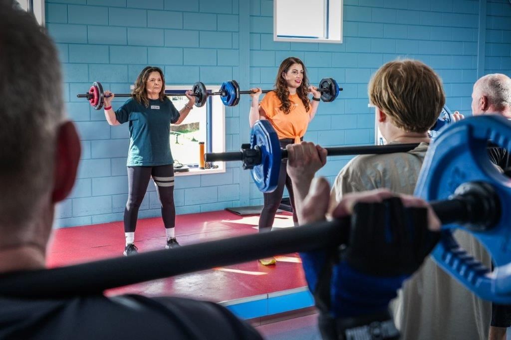 Fitness class in a blue gym, with people lifting barbells under an instructor's guidance
