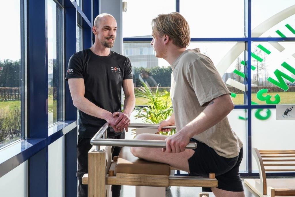 Een man helpt een andere man bij het gebruik van fitnessapparatuur in een kamer met een groot raam.