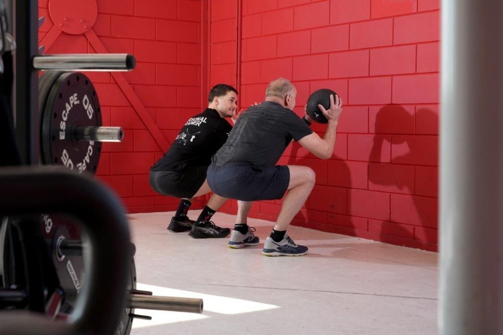 Twee mannen doen wall squats met een medicijnbal in een sportschool met rode muren.