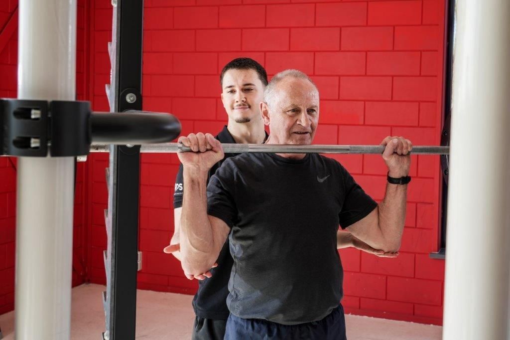 Een trainer ziet een oudere man squats doen met een halter in een sportschool met rode muren.