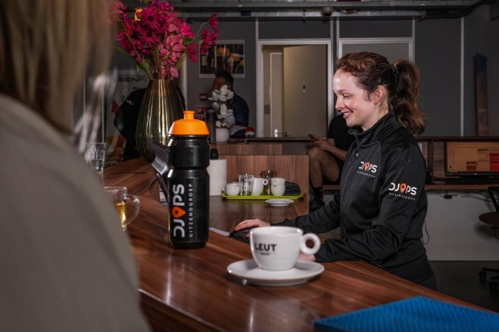 Vrouw aan de balie, lachend, met een kopje koffie en een fles water, in een sportschool of studio.