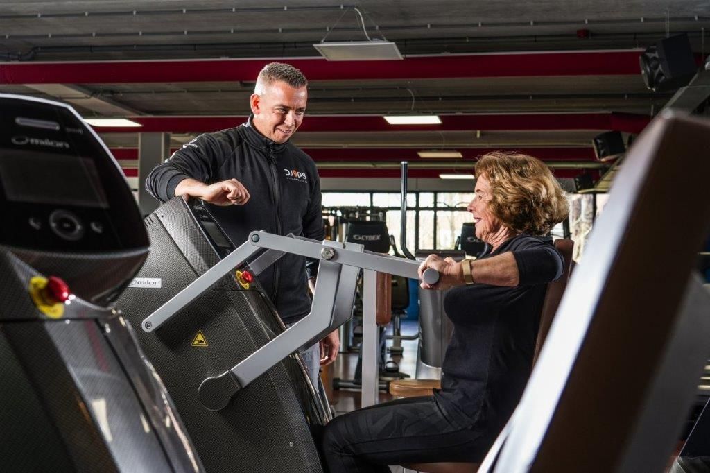 Man helpt vrouw met het gebruiken van een borstpersmachine in de sportschool.