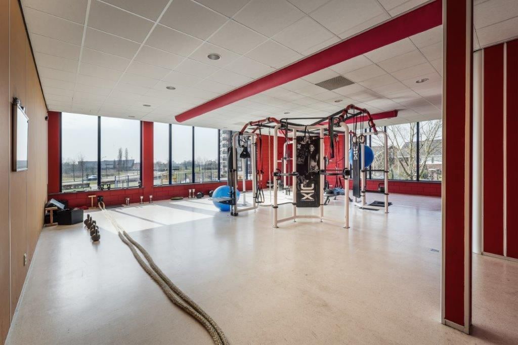 Sportschool met fitnessapparatuur, grote ramen, rode accenten en beige vloer.
