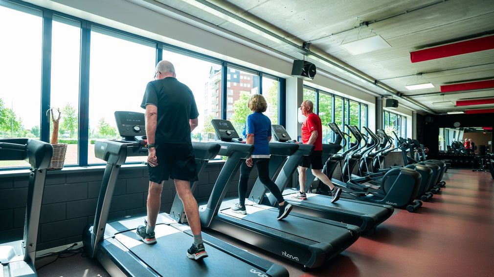 Man en vrouw in de sportschool, op crosstrainers. Man lacht, vrouw fitnesst. De setting is een fitnesscentrum.