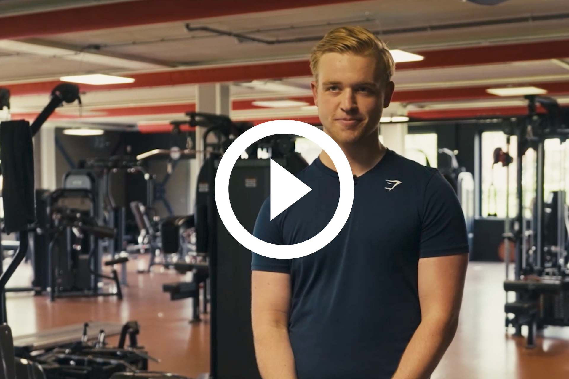 Een man staat in een sportschool, gekleed in een donker shirt, met fitnessapparatuur op de achtergrond.