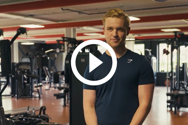 Een man staat in een sportschool, gekleed in een donker shirt, met fitnessapparatuur op de achtergrond.