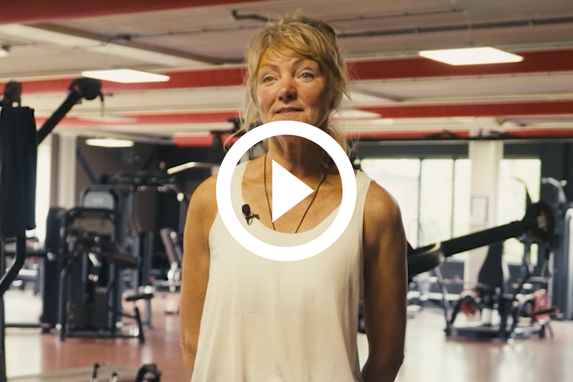 Vrouw staat in een sportschool en lacht naar de camera, met fitnessapparatuur op de achtergrond.