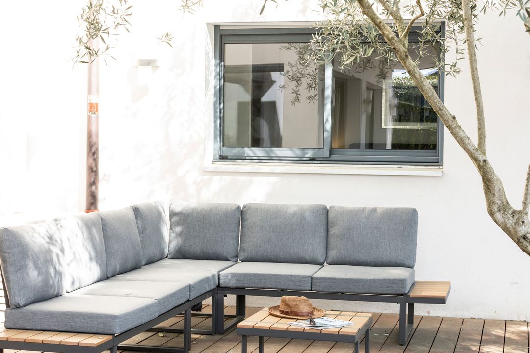 Modern patio seating with a gray sectional, wooden coffee table, and white wall beside a window