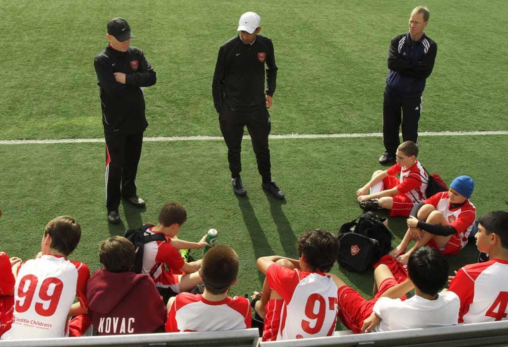 Soccer coaches in black stand on the field, speaking to players in red and white seated on the sideline.