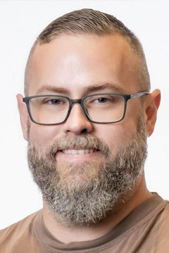 Portrait of a bearded man wearing glasses and a brown shirt, smiling against a light background