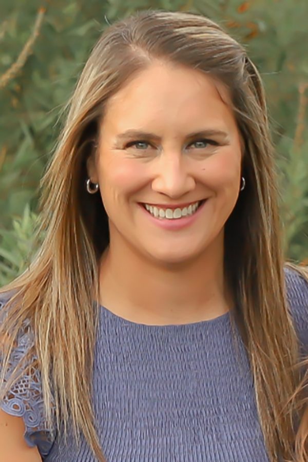 Smiling person in a blue top with long hair, standing outdoors against a blurred green background
