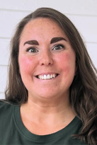 Smiling woman with long brown hair wearing a dark green shirt against a white brick wall