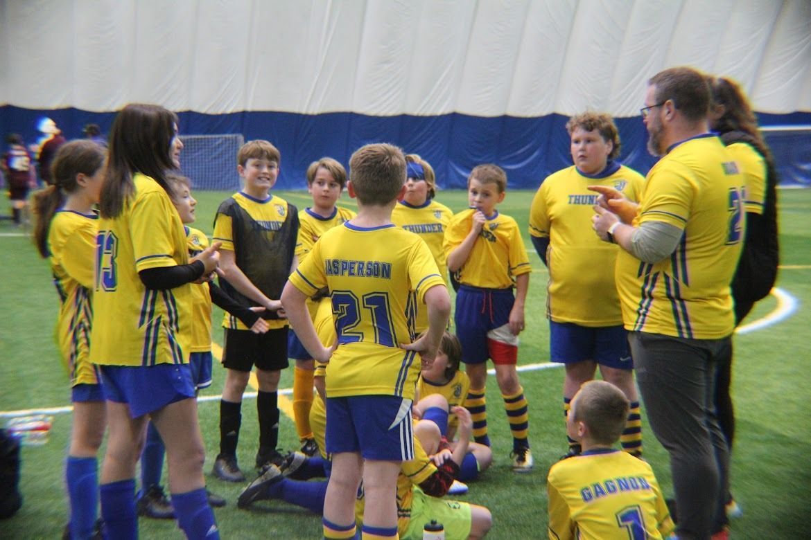 Youth soccer team in yellow jerseys huddles indoors on turf during a break.