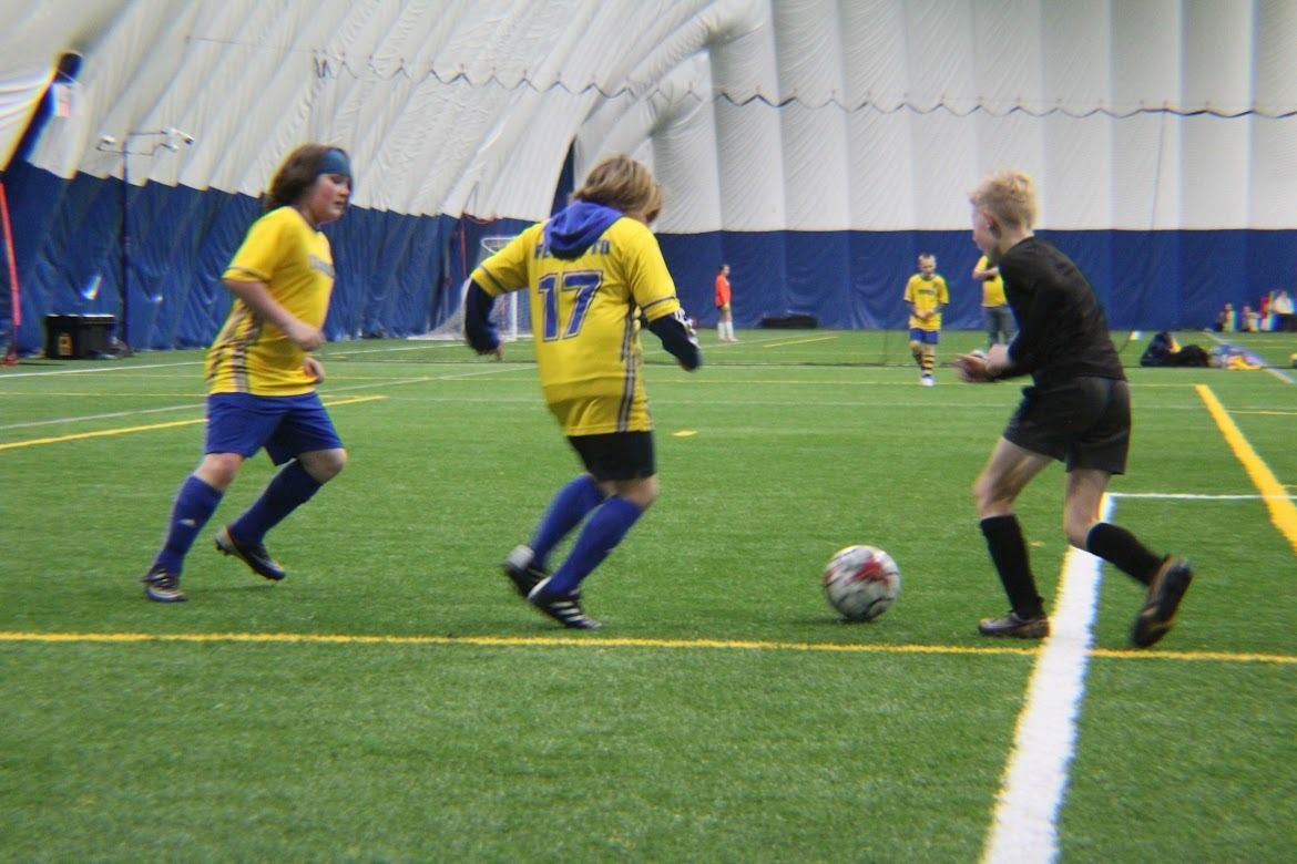 Youth soccer players in yellow and black jerseys chase the ball on an indoor turf field.