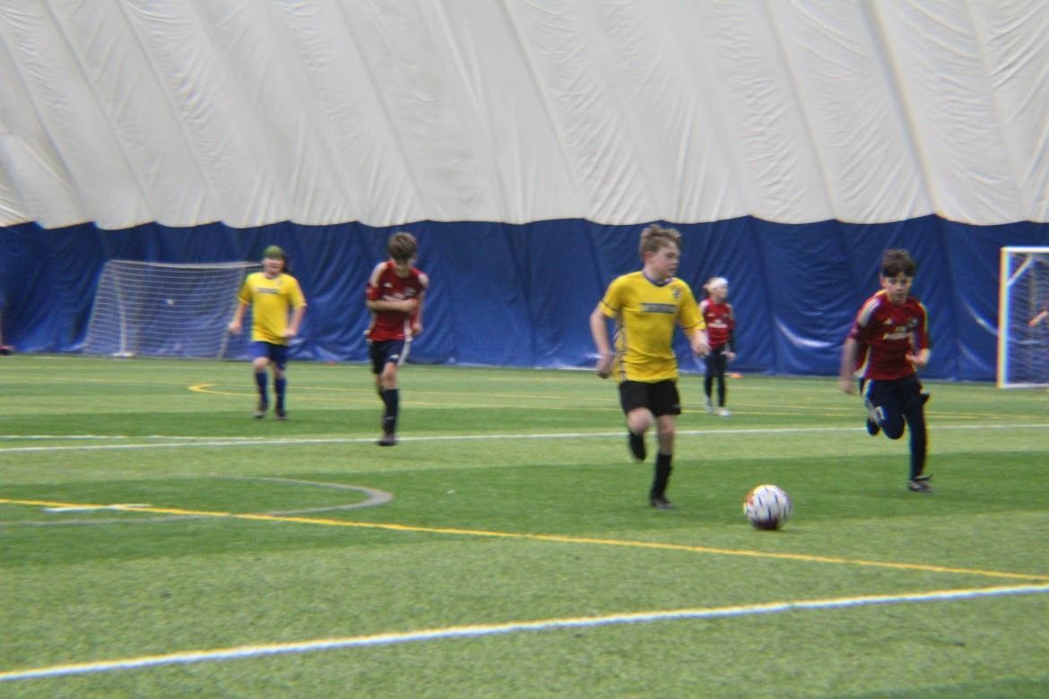 Kids playing soccer indoors on green turf, with yellow and red jerseys chasing the ball near the goal