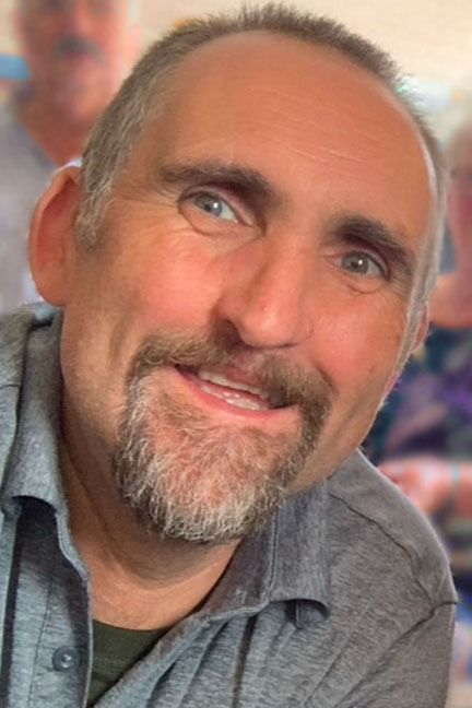 Close-up of a smiling man with a gray beard and blue eyes, wearing a gray shirt against a blurred background