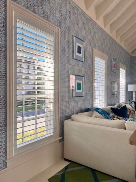 Bright living room with patterned wallpaper, white shutters, and a cream sofa by the windows.
