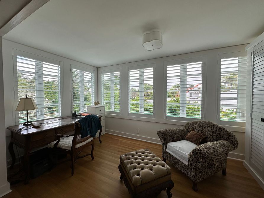 Bright sitting room with large windows, desk, armchair, and tufted ottoman; white shutters and greenery outside.