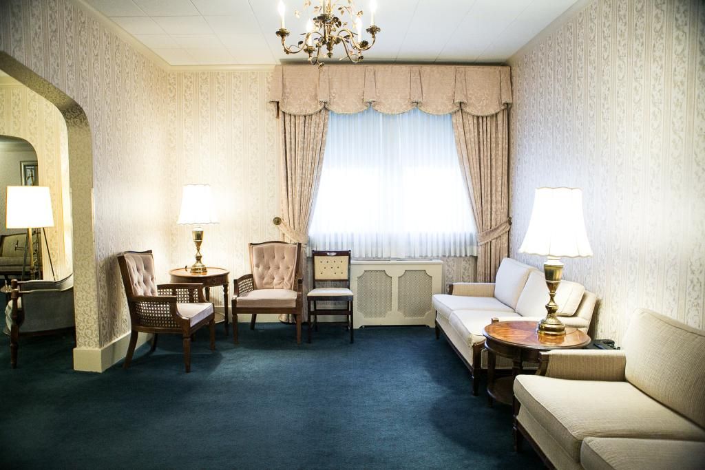 Living room with blue carpet, light walls, and floral window curtains. Sofas, chairs, and lamps present.