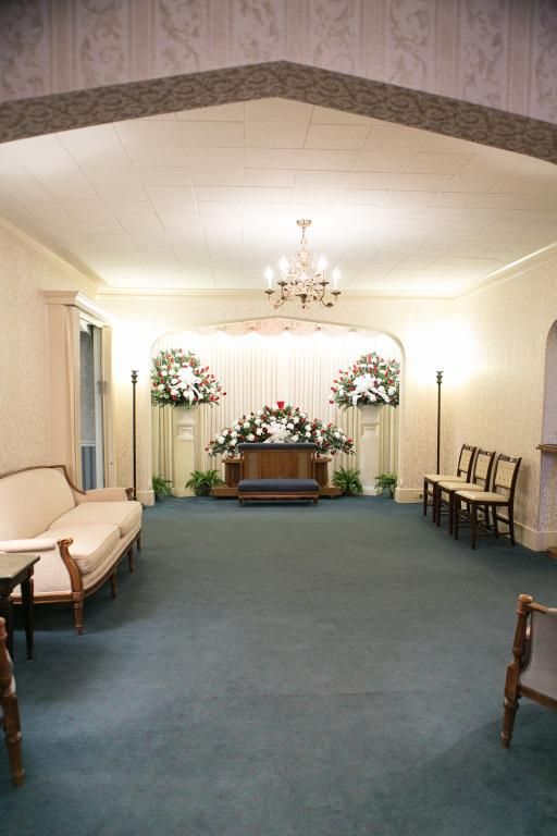 Funeral home chapel with blue carpet, cream walls, flowers, and seating.