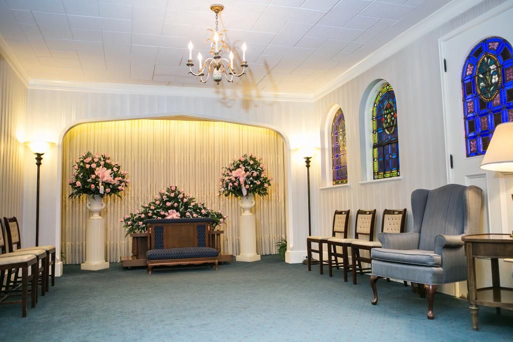 Funeral home interior, with a casket, floral arrangements, and stained-glass windows.